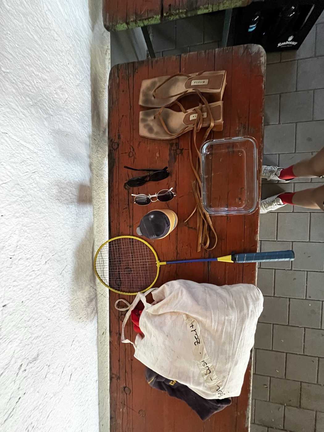 Fundstücke auf einer Holzbank: Sandalen, Sonnenbrillen, Trinkflasche, Federballschläger, Beutel, Tupperdose